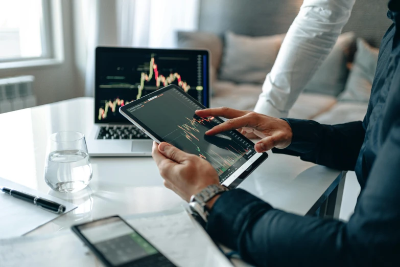 a couple of people sitting at a table with laptops, trending on pexels, analytical art, displaying stock charts, using a magical tablet, thumbnail, holding close