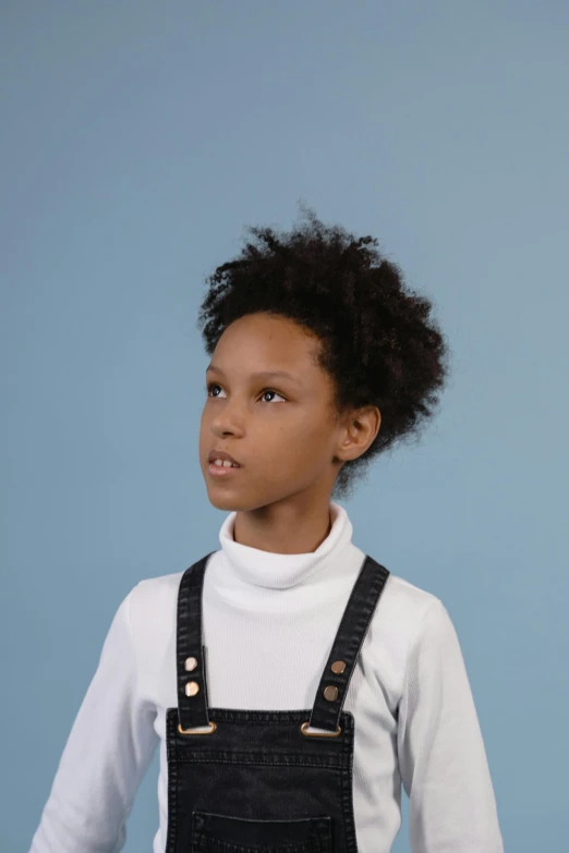 a little girl standing in front of a blue wall, an album cover, inspired by Theo Constanté, trending on unsplash, afrofuturism, in white turtleneck shirt, wearing overalls, portrait. 8 k high definition, cinematic still