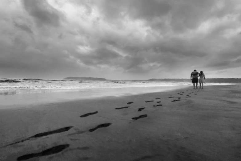 two people walking on a beach with footprints in the sand, a black and white photo, by Felix-Kelly, pembrokeshire, rainy, heavens, full width
