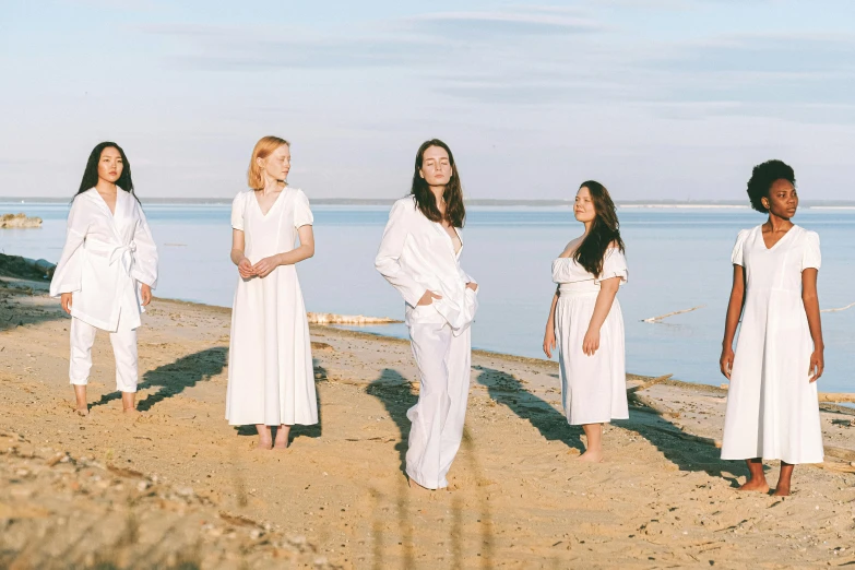 a group of women standing on top of a sandy beach, an album cover, unsplash, antipodeans, wearing white pajamas, dressed in white robes, yulia nevskaya, band promo photo