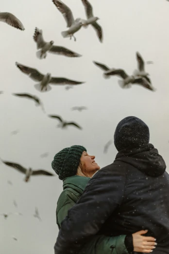 a man and a woman standing in front of a flock of birds, by Roar Kjernstad, pexels contest winner, snowing outside, seagull wearing luigis hat, touching heads, overcast skies