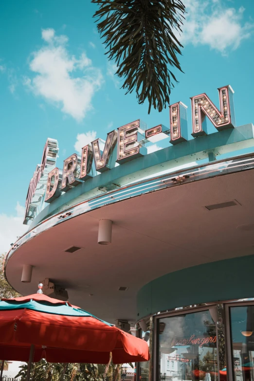 a building with a sign and umbrellas in front of it, poster art, by Dan Luvisi, trending on unsplash, retrofuturism, drive in movie theater, miami beach, 5 0's diner, bumper cars