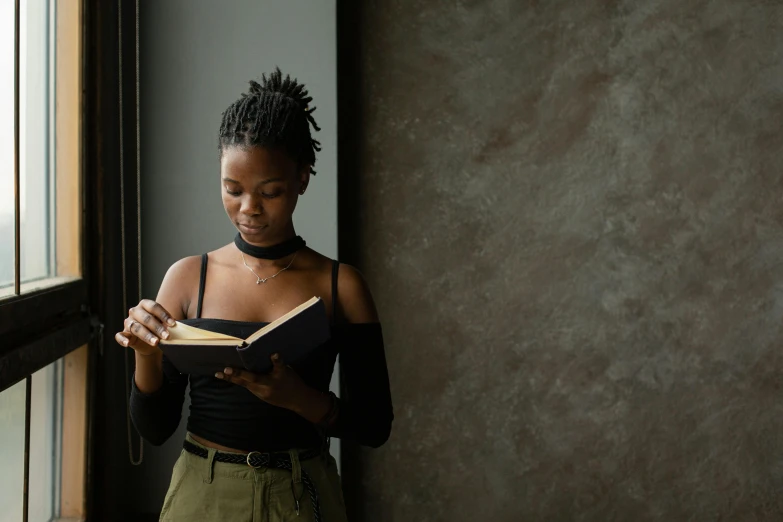 a woman standing by a window reading a book, a portrait, by Lily Delissa Joseph, pexels contest winner, black young woman, girl in studio, holding notebook, a black choker