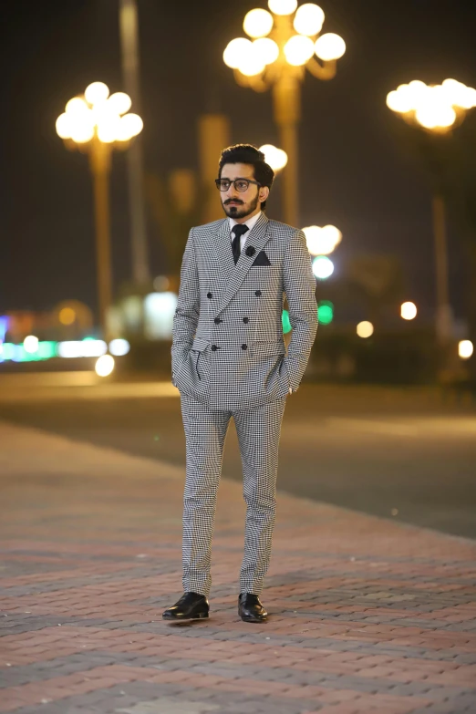 a man in a suit standing on a sidewalk, inspired by Ahmed Karahisari, jeddah city street, grey suit, during night, wearing a suit and glasses