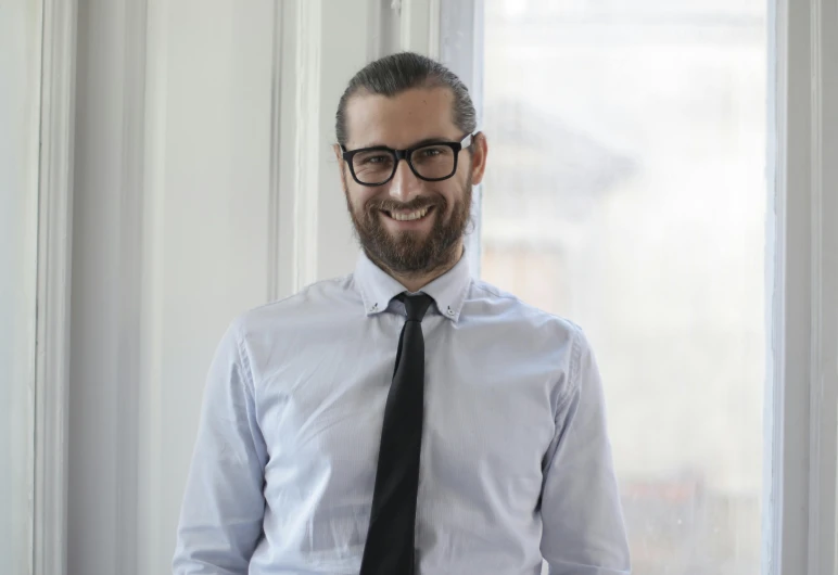 a man in a dress shirt and tie standing in front of a window, by Patrick Pietropoli, wearing black rimmed glasses, hasan piker, 2019 trending photo, smiling male