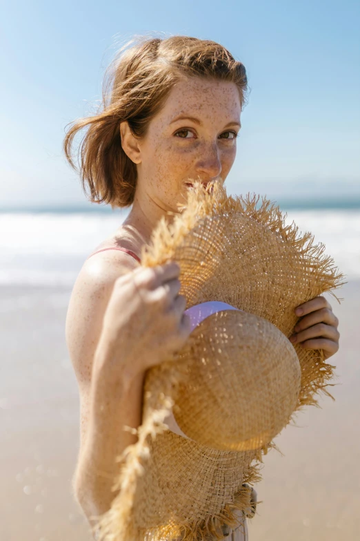 a woman standing on a beach holding a straw hat, a portrait, pexels contest winner, renaissance, her face flushing and sweat, sun drenched, freckled pale skin, round-cropped