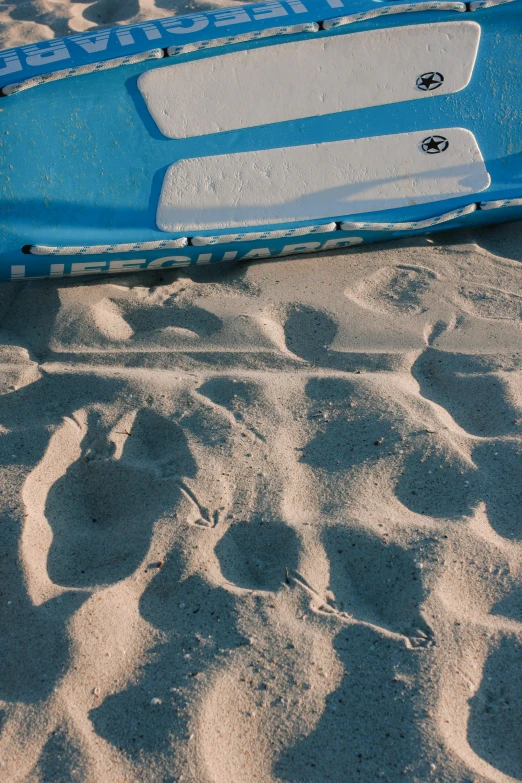 a blue surfboard sitting on top of a sandy beach, footprints in the sand, shadowy cat under the bed', boat, up-close