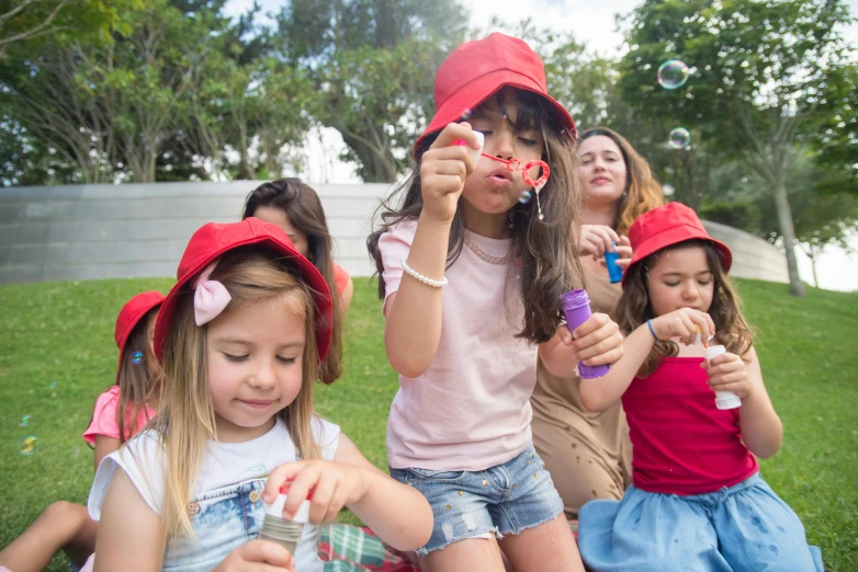 a group of young girls sitting on top of a lush green field, by Lilia Alvarado, pexels contest winner, pop art, red cap with a capital m, soap bubbles, at a birthday party, in sao paulo