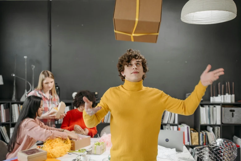 a group of people sitting around a table, yellow clothes, things hanging from ceiling, creative coder with a computer, holding gift