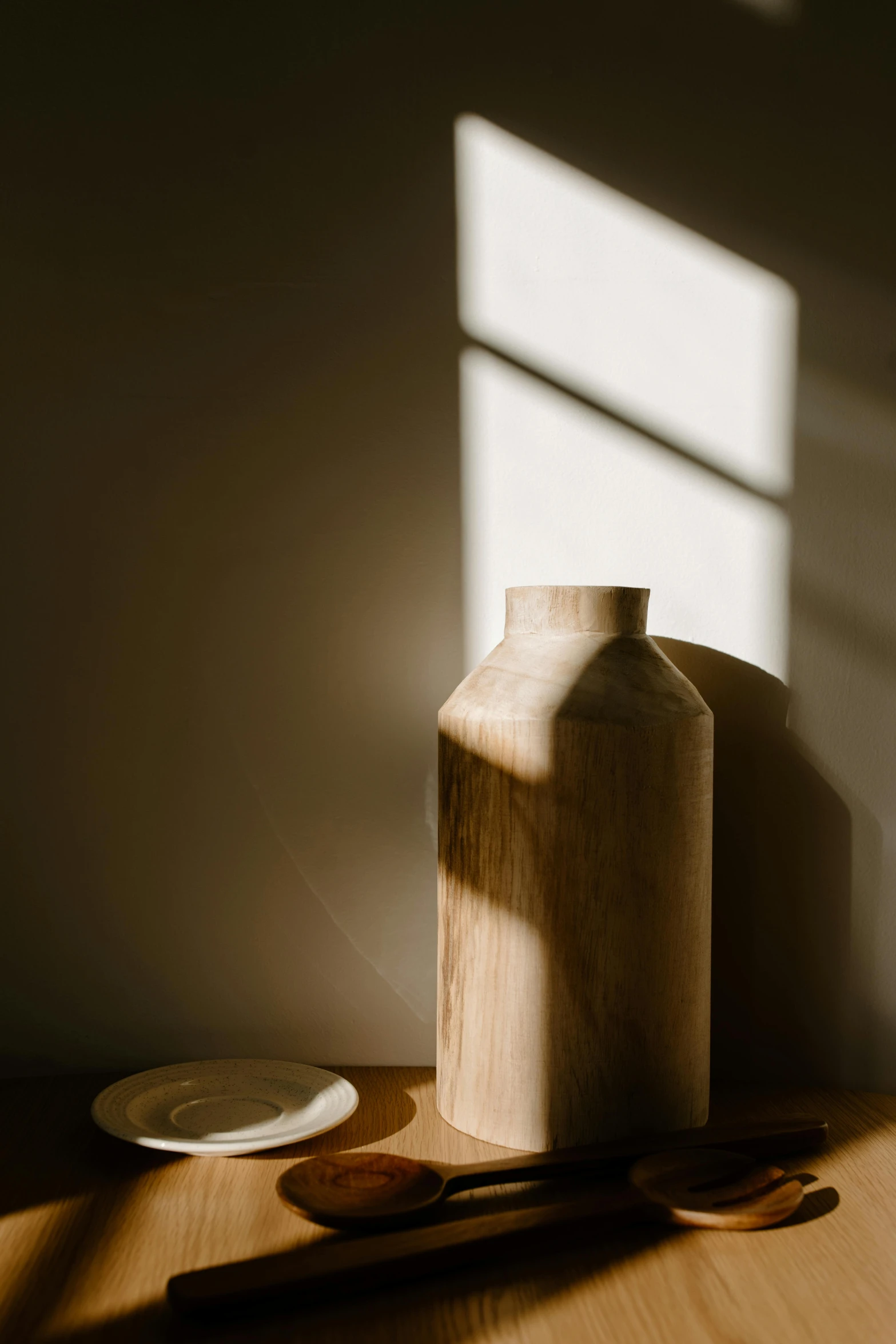 a vase sitting on top of a wooden table next to a plate, unsplash, light and space, soft light - n 9, backlighted, large tall, sun lit