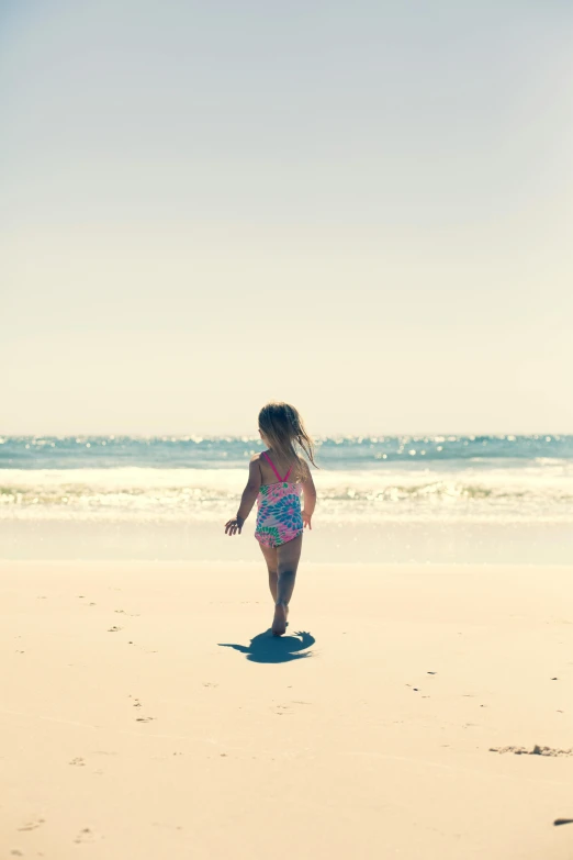 a little girl standing on top of a sandy beach, walking away from the camera, australian beach, sun - drenched