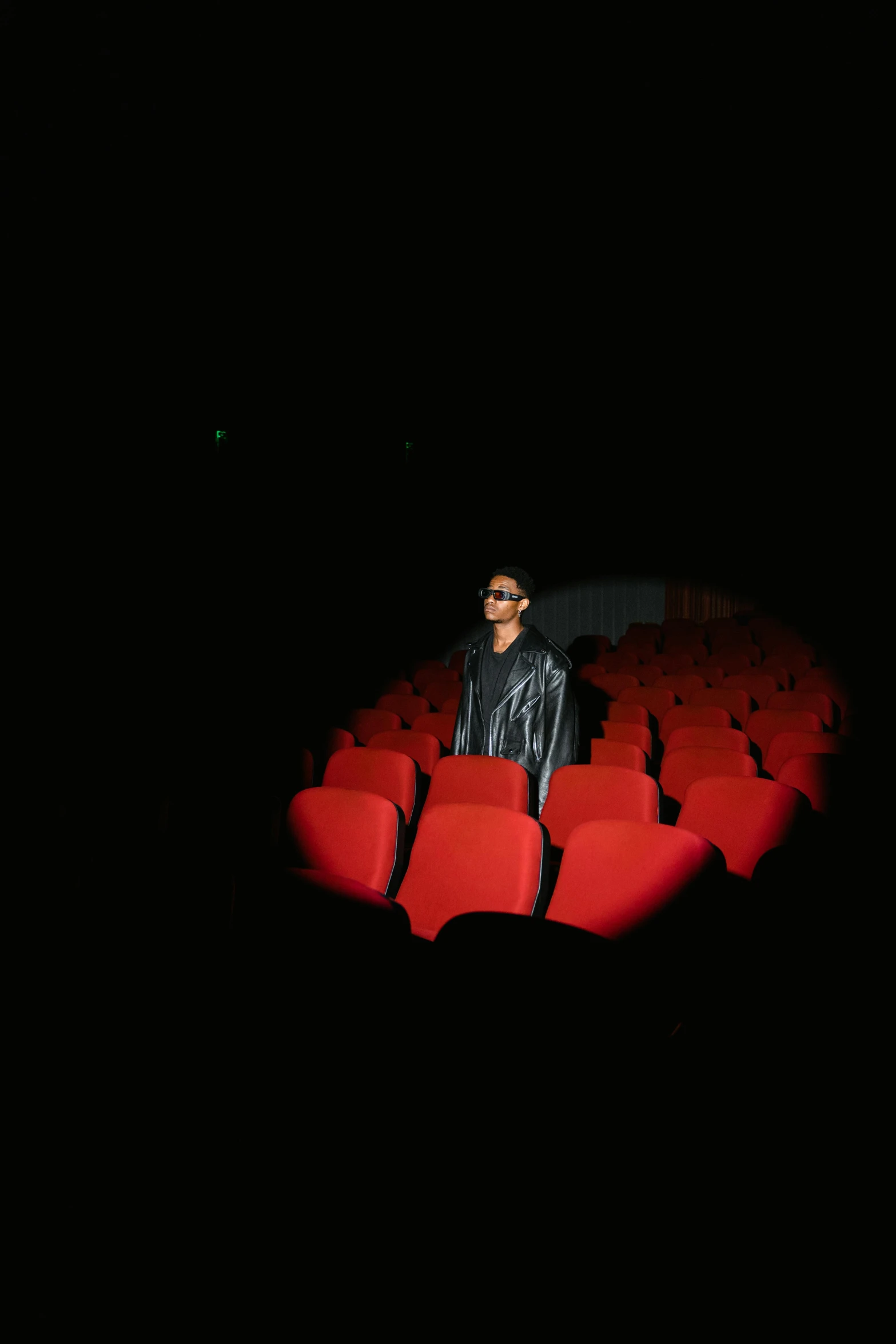 a man standing in front of a row of red chairs, an album cover, pexels, sitting in a movie theater, arca album cover, [ theatrical ], with a black background