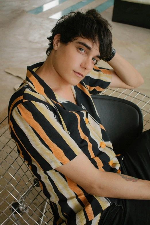 a young man sitting on top of a metal chair, by Robbie Trevino, trending on pexels, renaissance, striped shirt, black and orange colour palette, loose - fitting blouses, androgynous face