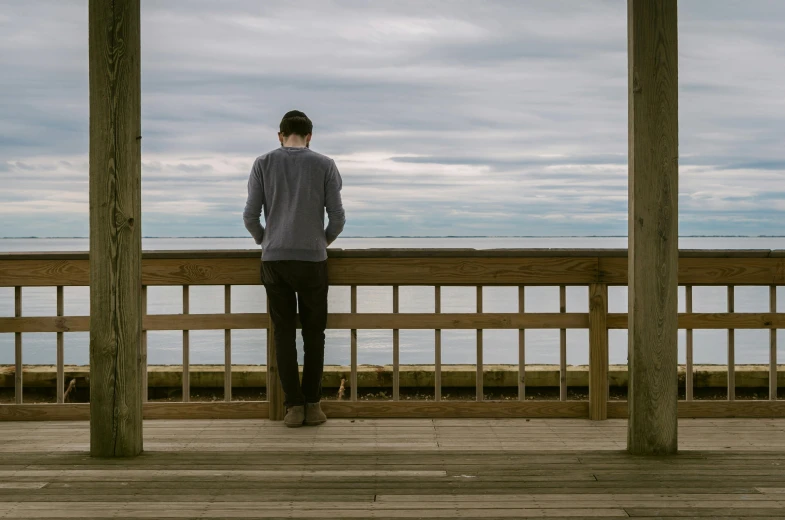 a man standing on a deck looking out at the ocean, a picture, unsplash, visual art, overcast day, she's sad, teenage boy, ignant