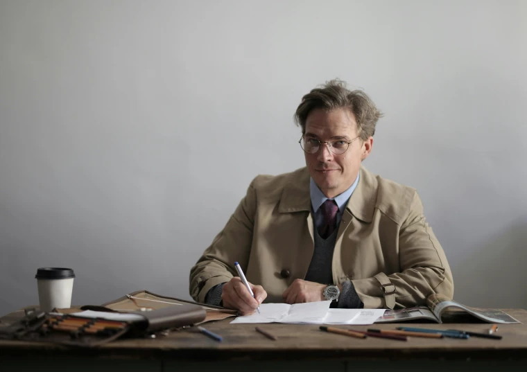 a man sitting at a table writing on a piece of paper, inspired by Alasdair Gray, press shot, professor clothes, kurt russell, ignant