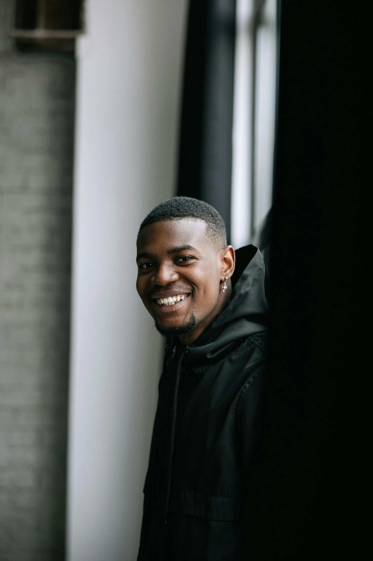 a man standing in front of a window smiling, in a black hoodie, black man, in front of white back drop, smiling sweetly