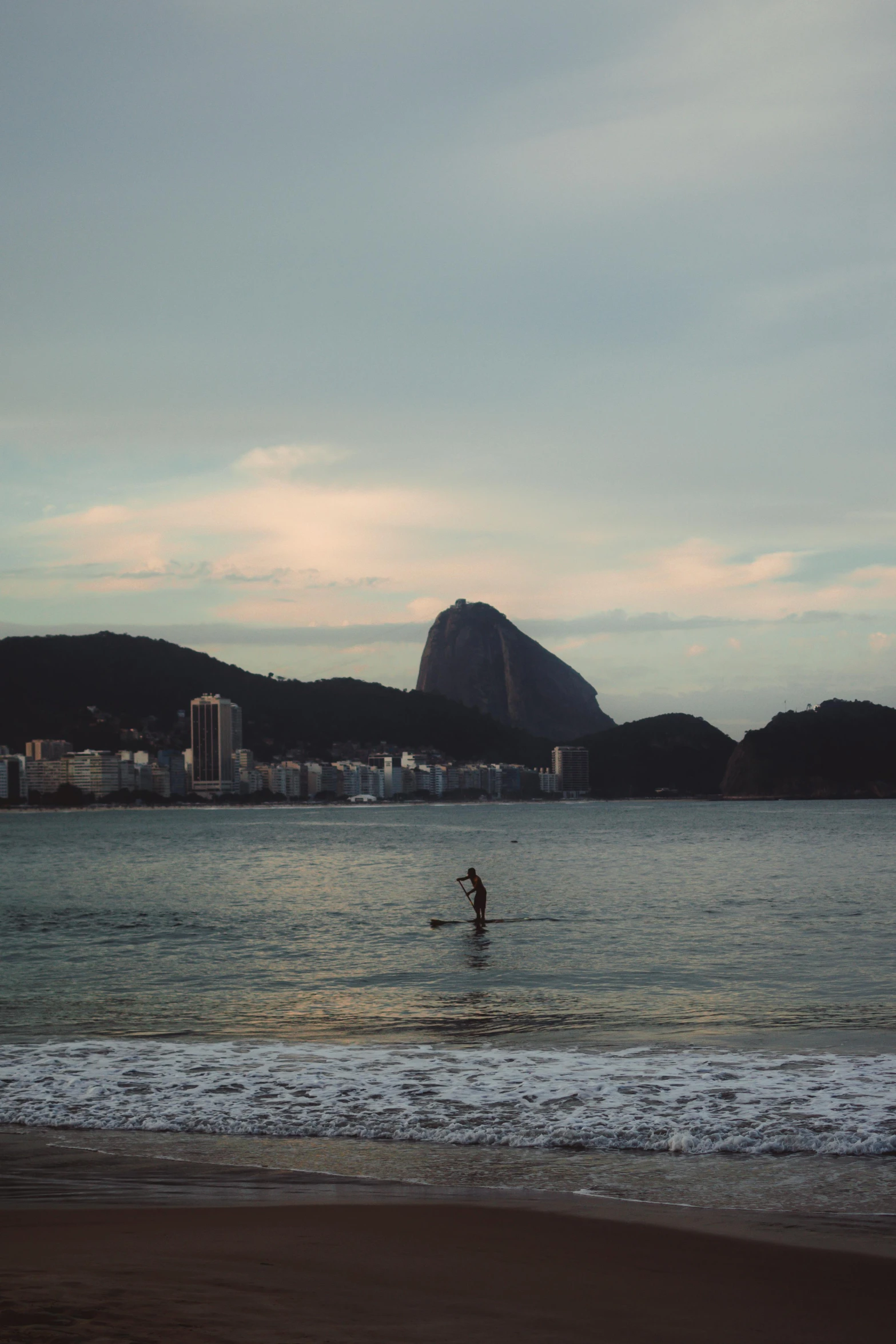 a man riding a surfboard on top of a sandy beach, inspired by Elsa Bleda, happening, cristo redentor, square, f / 1. 8, city view