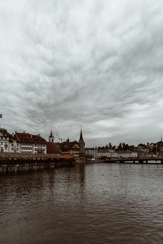 a body of water surrounded by buildings under a cloudy sky, inspired by Karl Stauffer-Bern, pexels contest winner, moody cloudy sky, low quality photo, brown, a quaint