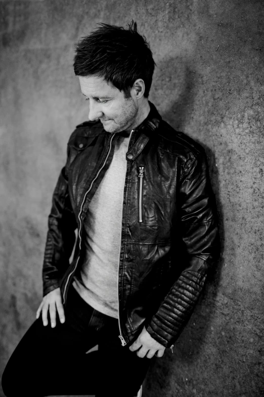 a man in a leather jacket leaning against a wall, an album cover, by Kevin Connor, pexels, jeremy renner, a handsome man，black short hair, paul rudd, back and white