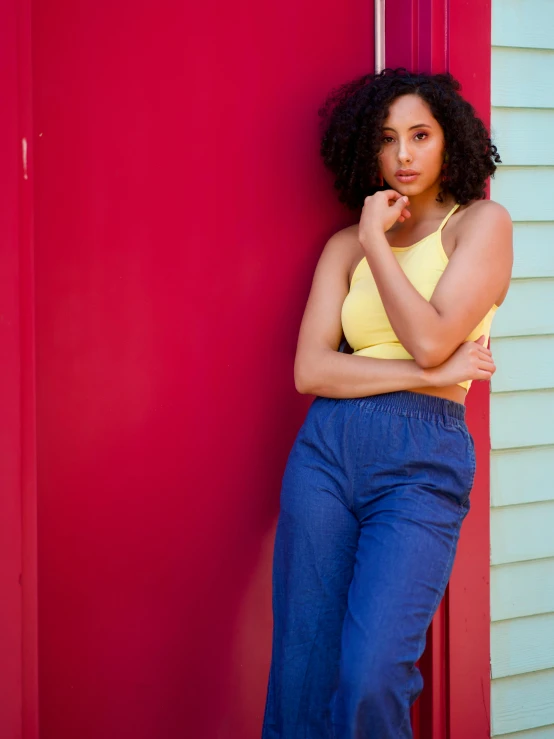 a woman leaning against a red door, by Arabella Rankin, pexels contest winner, red tank top and wide blue pants, wavy hair yellow theme, portrait of vanessa morgan, vintage clothing