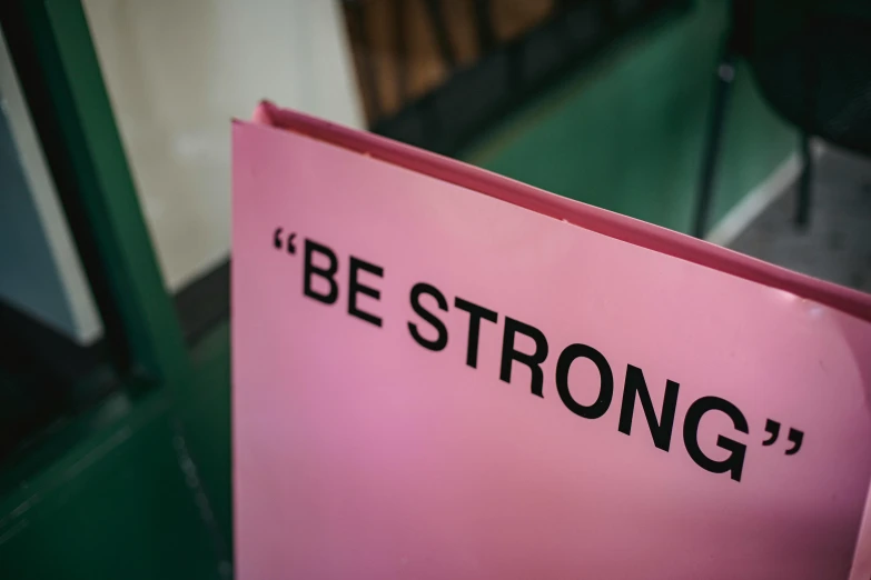 a pink sign with the words be strong written on it, a poster, trending on unsplash, bottom body close up, a book, strength, ( 3 1