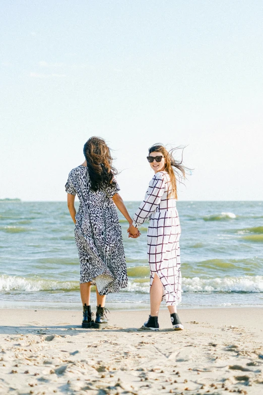 a couple of women standing on top of a sandy beach, standing next to water, profile image, patterned clothing, trending photo