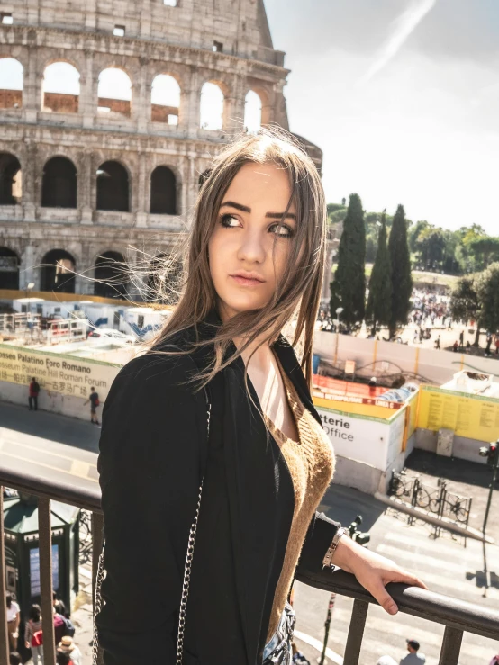 a woman posing for a picture in front of the colossion, by Julia Pishtar, coliseum backdrop, looking off to the side, 19-year-old girl, wearing a black jacket