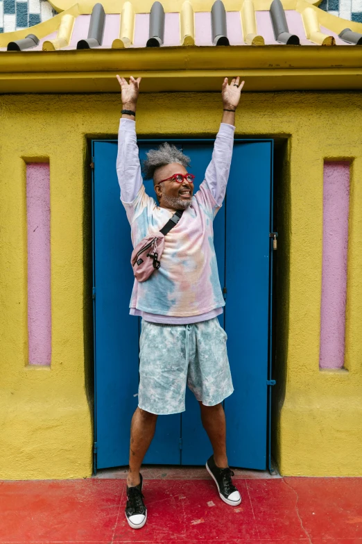 a man standing in front of a yellow building, maximalism, pose(arms up + happy), tie-dye, an oldman, hung above the door