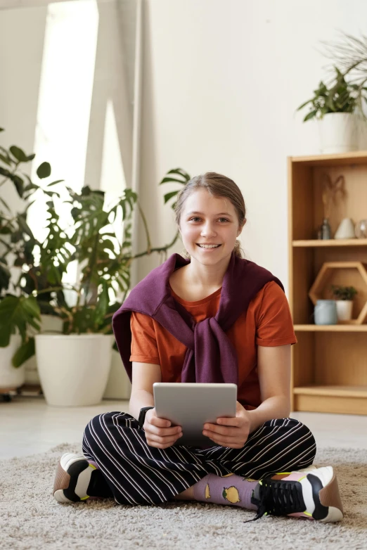 a woman sitting on the floor using a tablet computer, greta thunberg smiling, professional grade, next to a plant, relaxing and smiling at camera