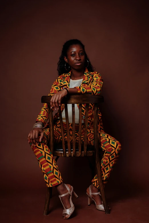 a woman sitting on top of a wooden chair, by Lily Delissa Joseph, pexels contest winner, afrofuturism, wearing a colorful men's suit, black teenage girl, symmetrical image, brown