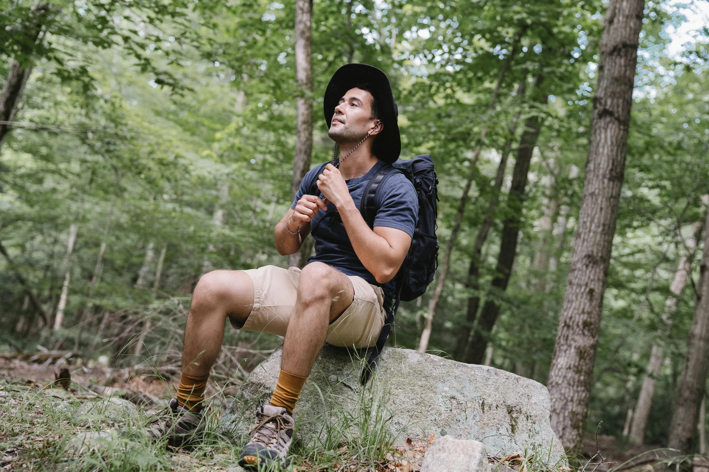 a man sitting on a rock in the woods, wearing a travel hat, wearing black shorts, nathan fielder, thumbnail