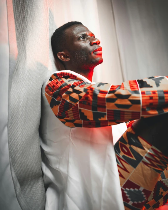 a man sitting on top of a bed next to a window, an album cover, inspired by David Bailly, pexels contest winner, afrofuturism, wearing an african dress, lgbtq, his arms are behind his back, looking confident