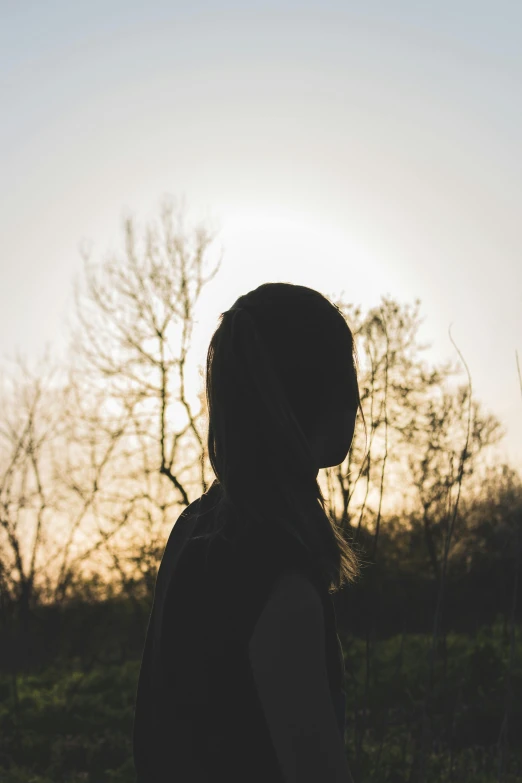 a silhouette of a person standing in a field, woman's face looking off camera, trees in the background, ((sunset)), pictured from the shoulders up