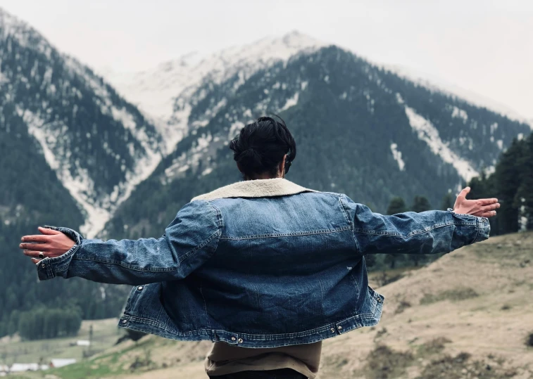 a man standing on top of a lush green hillside, an album cover, pexels contest winner, wearing a jeans jackets, his arms spread. ready to fly, himalayas, timothee chalamet