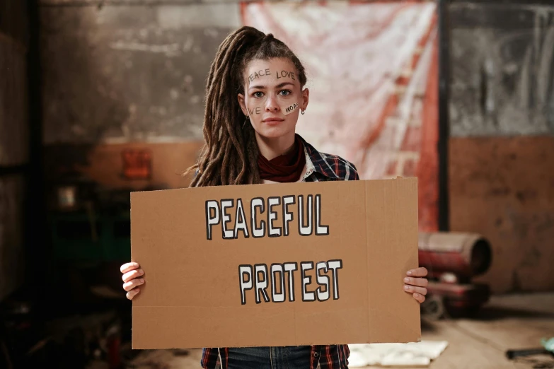 a woman holding a sign that says peaceful protest, an album cover, trending on pexels, dreads, natalia dyer, artem chebokha, cardboard