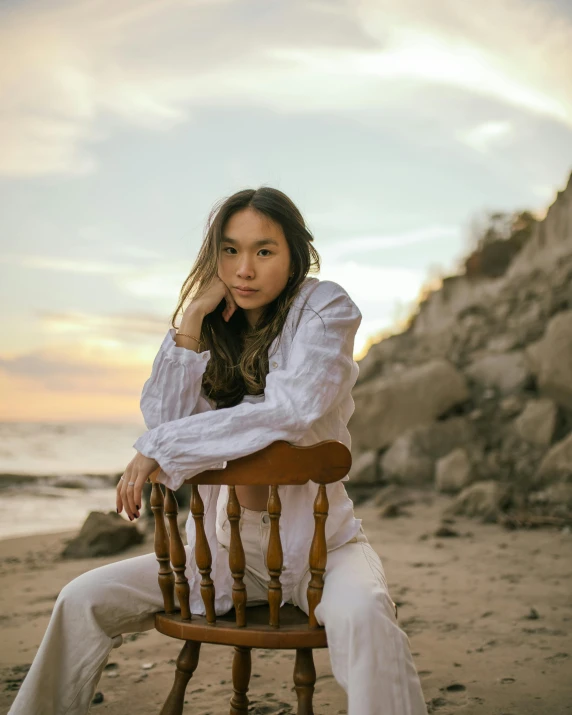 a woman sitting on top of a wooden chair on a beach, inspired by helen huang, unsplash, romanticism, in white turtleneck shirt, looking straight to the camera, queer woman, ashteroth