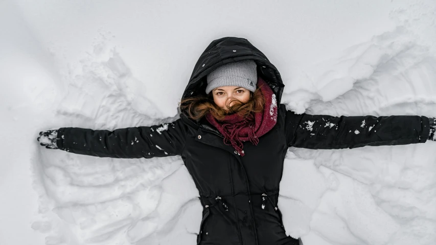 a woman making a snow angel in the snow, pexels contest winner, looking up at camera, black, puffer jacket, smiling down from above