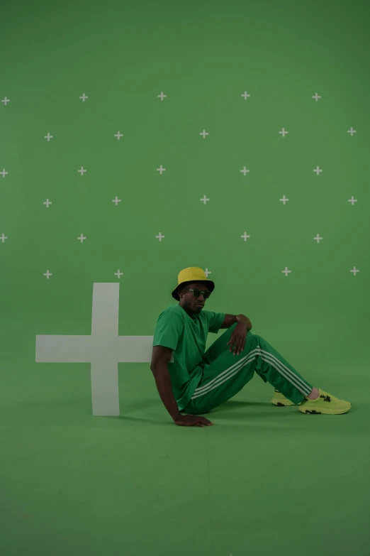 a man sitting on the ground in front of a cross, an album cover, trending on pexels, green hat, wearing track and field suit, ( ( dark skin ) ), vfx shot