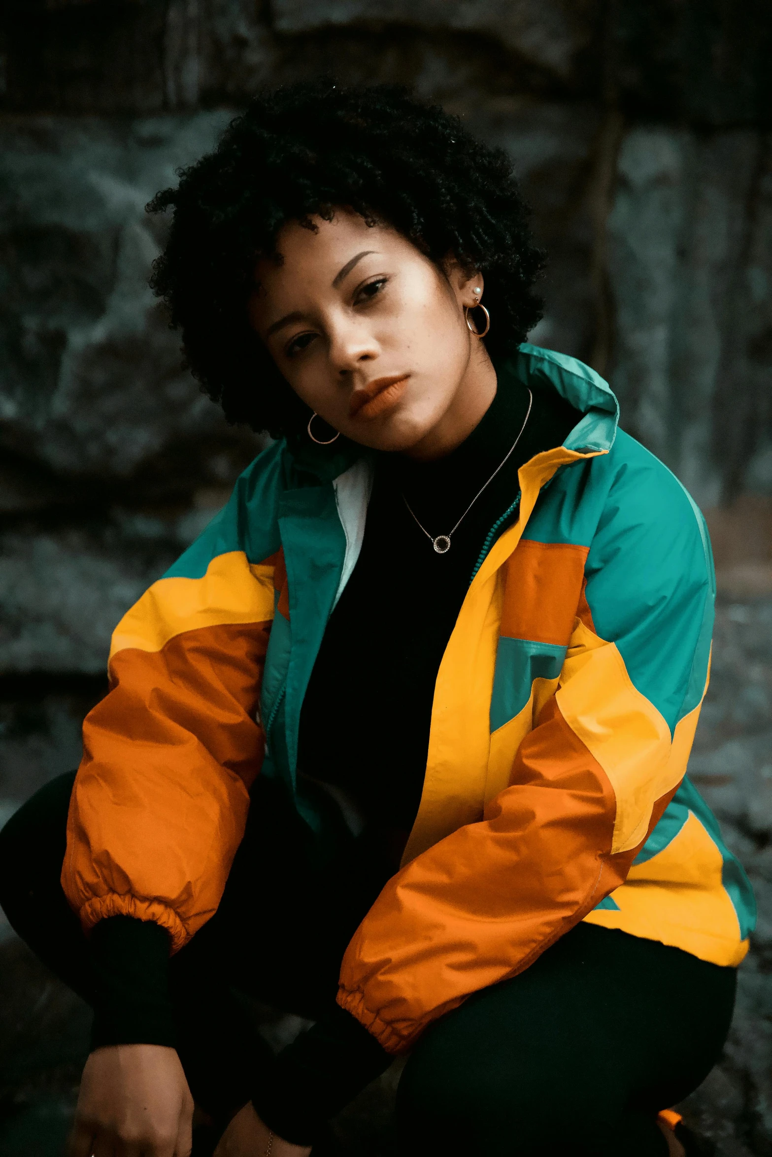 a woman in an orange and green jacket sitting on a rock, trending on pexels, mixed race, streetwear, black and yellow, teal