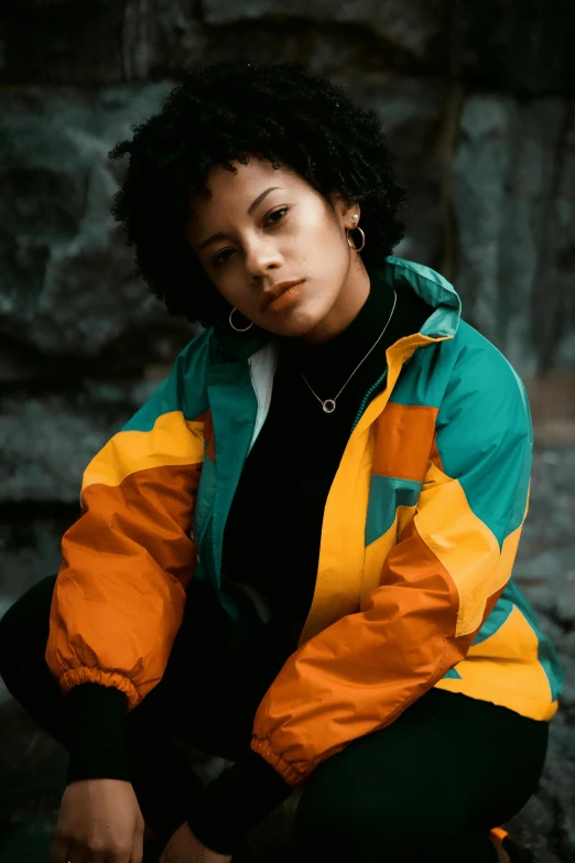 a woman in an orange and green jacket sitting on a rock, trending on pexels, mixed race, streetwear, black and yellow, teal