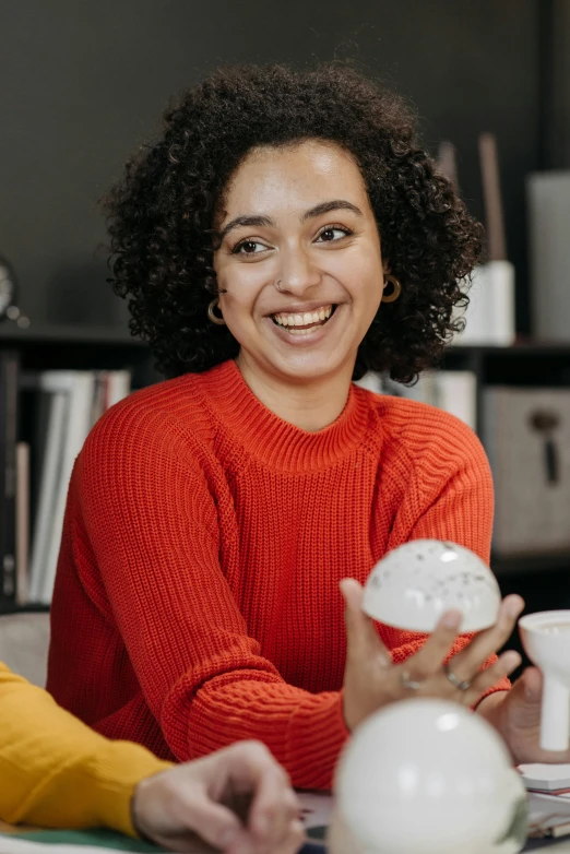 a couple of people that are sitting at a table, a woman holding an orb, dark short curly hair smiling, wearing casual sweater, promo image