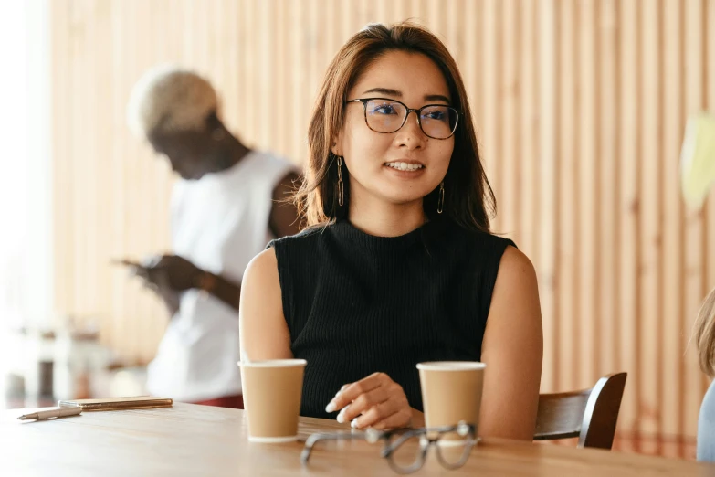 a woman sitting at a table with a cup of coffee, a portrait, trending on pexels, zeen chin and terada katsuya, wearing small round glasses, aussie baristas, female in office dress