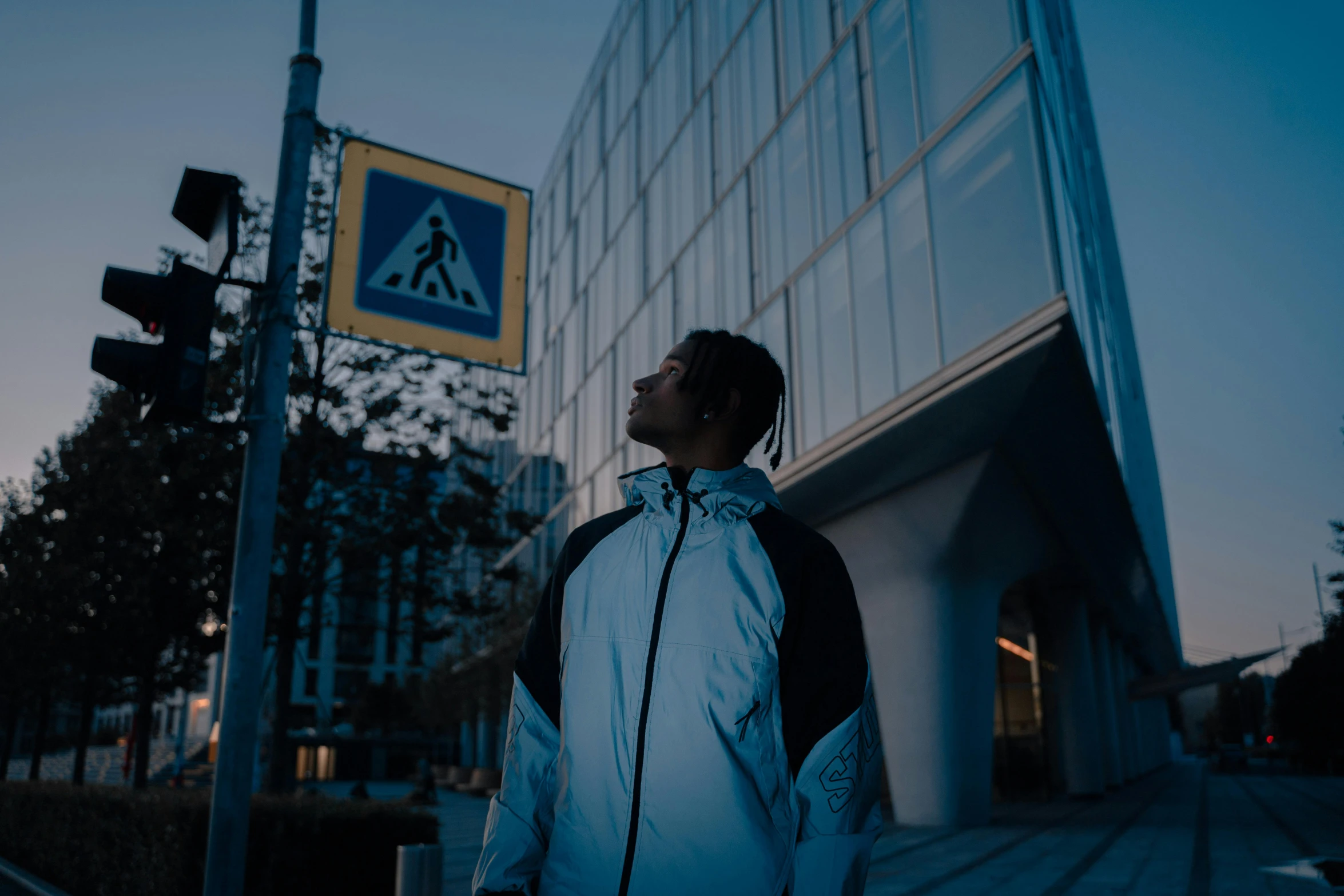 a man standing in front of a tall building, an album cover, unsplash, chappie in an adidas track suit, night time footage, with backlight, reflective puffer jacket