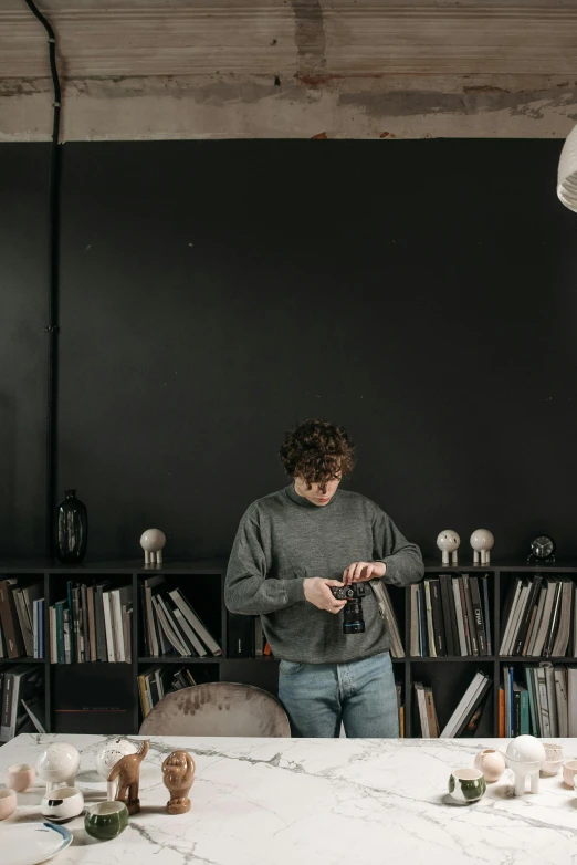 a man that is standing in front of a table, pexels contest winner, bookshelf, darkslategray wall, checking her phone, grey sweater