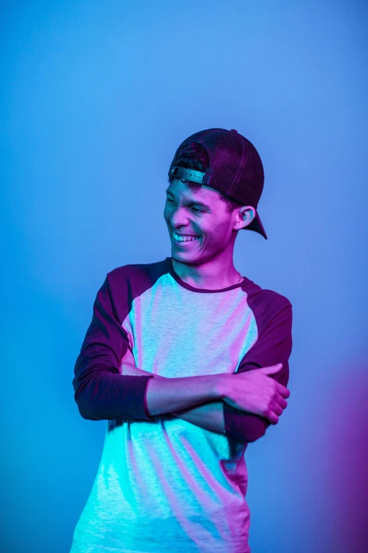 a man standing in front of a blue background, an album cover, inspired by John Luke, pexels contest winner, graffiti, smileing nright, he is wearing a hat, dark. no text, bisexual lighting