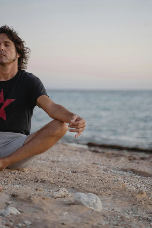 a man sitting on top of a sandy beach next to the ocean, a portrait, pexels, black shirt with red star, yoga, avatar image, mediterranean