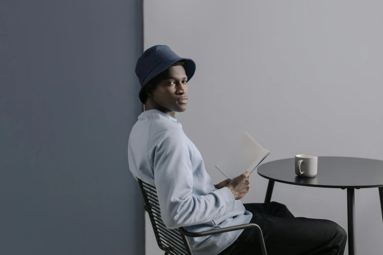 a woman sitting at a table reading a book, a character portrait, pexels contest winner, he is wearing a hat, black teenage boy, sitting on designer chair, promotional image