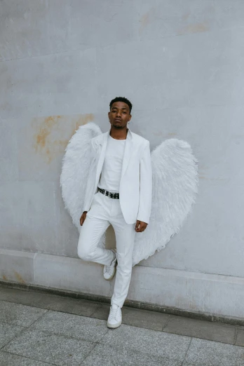 a man in a white suit leaning against a wall, an album cover, inspired by Louis Hersent, pexels contest winner, big angel wings wide open, black teenage boy, cupid, comme des garcon campaign