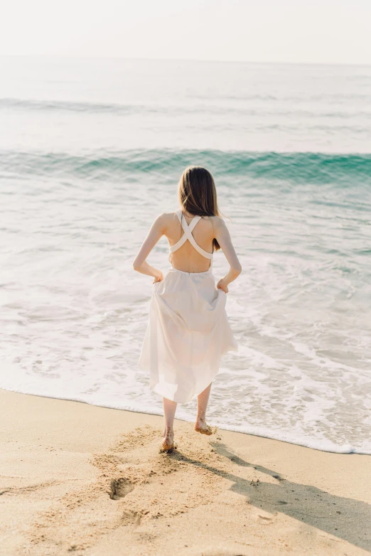 a woman standing on top of a beach next to the ocean, open back dress, wavy, wearing white cloths, about to step on you