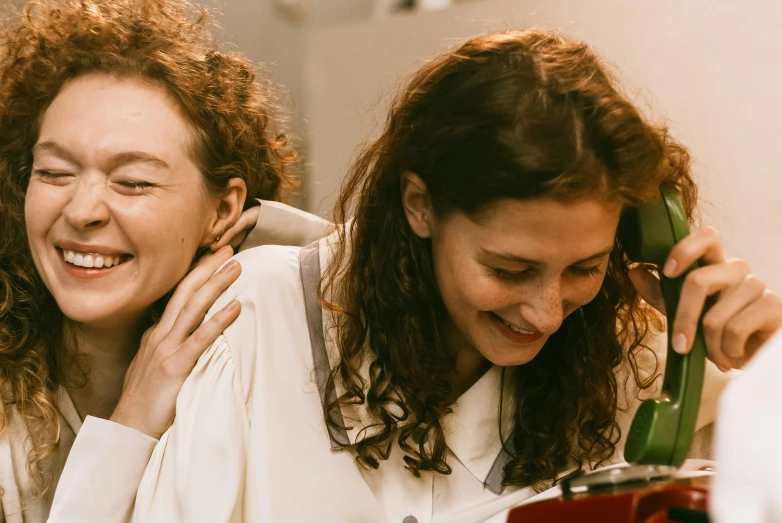 a woman talking on a phone next to another woman, trending on pexels, happening, dark short curly hair smiling, wearing white robes, lorde, [[[[grinning evily]]]]